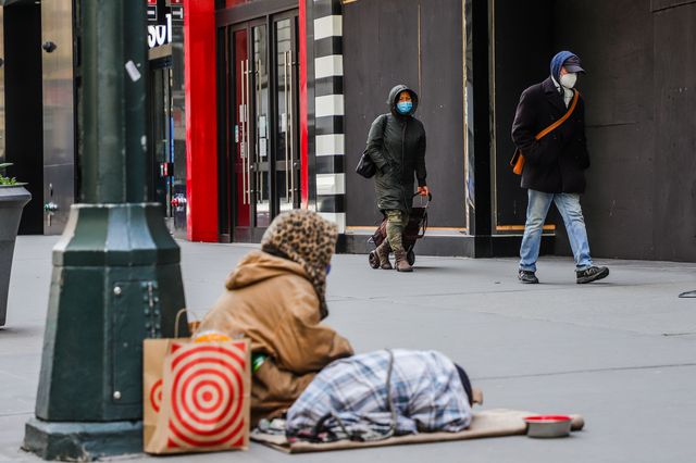 A homeless person, with their back to the camera, sits on a Midtown sidewalk while two people wearing masks walk by